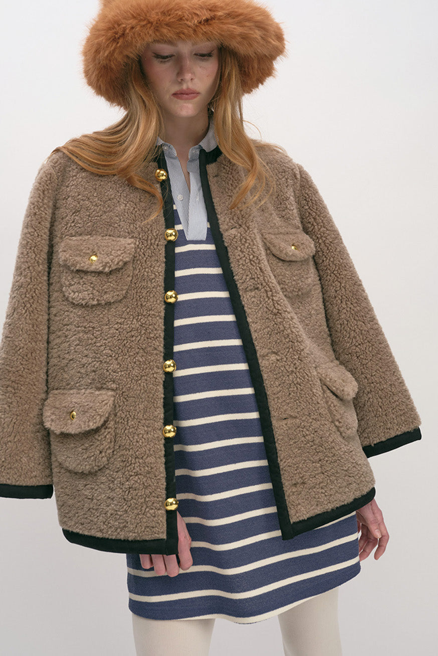 Featuring a collarless neckline, contrast trim detailing, and a front button closure with gold-tone buttons, the jacket blends functional design with refined accents.